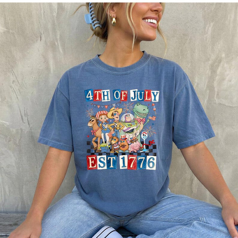 May include: Blue t-shirt with a graphic featuring cartoon characters and the text "4TH OF JULY EST 1776" in red, white, and blue. The shirt has a vintage, faded appearance.