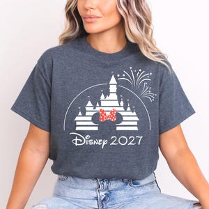 May include: Dark gray t-shirt featuring a white graphic of a castle with a red and white polka dot bow, fireworks, and the text "Disney 2027". The shirt is a crew neck.