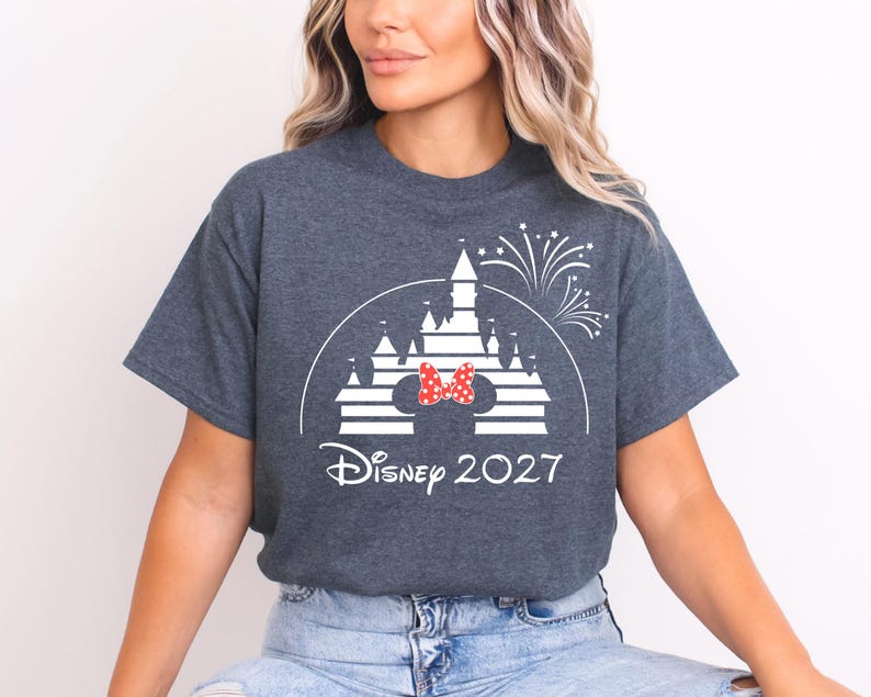 May include: Dark gray t-shirt featuring a white graphic of a castle with a red and white polka dot bow, fireworks, and the text "Disney 2027". The shirt is a crew neck.