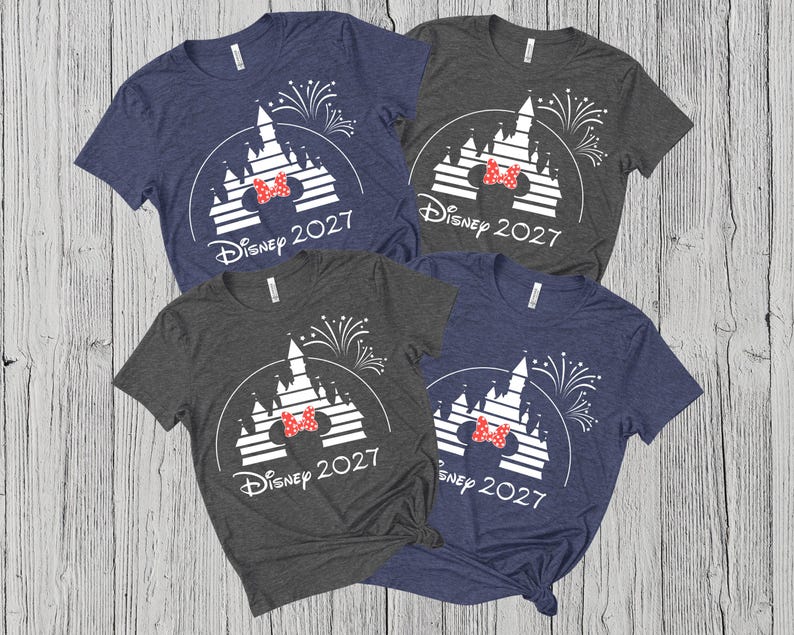 May include: Four short-sleeved t-shirts in navy blue and gray, each featuring a white castle design with a red bow and the text "Disney 2027". The shirts are arranged on a light wood-grain surface.