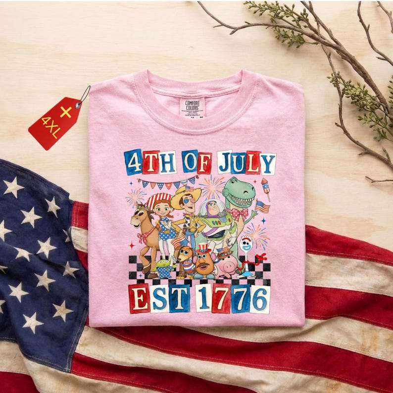 May include: A pink Comfort Colors t-shirt with a graphic of animated characters celebrating the 4th of July. The text "4TH OF JULY" and "EST 1776" are displayed. A red tag with "4XL" is attached.