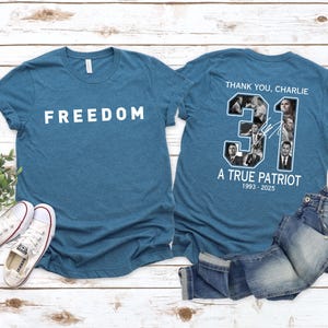 K&ouml;nnte beinhalten: T&uuml;rkisfarbene T-Shirts mit wei&szlig;em Text, der "FREEDOM" und "THANK YOU, CHARLIE" lautet. Auf der R&uuml;ckseite des Shirts befindet sich eine Collage von Bildern und der Text "31 A TRUE PATRIOT 1993-2025". Ein Paar wei&szlig;e Turnschuhe und blaue Jeans sind ebenfalls im Bild.