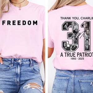 K&ouml;nnte beinhalten: Ein hellrosa T-Shirt mit dem Wort "FREEDOM" in fetten schwarzen Buchstaben auf der Vorderseite. Auf der R&uuml;ckseite des T-Shirts steht der Text "THANK YOU, CHARLIE" &uuml;ber der Zahl "31" mit Bildern und dem Text "A TRUE PATRIOT 1993-2025".