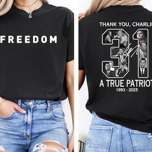 K&ouml;nnte beinhalten: Schwarzes T-Shirt mit dem Wort "FREEDOM" in wei&szlig;en Blockbuchstaben auf der Vorderseite. Auf der R&uuml;ckseite des Shirts steht der Text "THANK YOU, CHARLIE" &uuml;ber der Nummer "31" mit Bildern und dem Text "A TRUE PATRIOT 1993-2025".