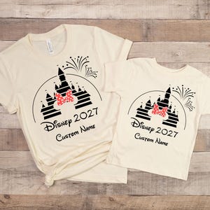 May include: Two cream-colored t-shirts with a black graphic of a castle, fireworks, and a red bow. The text "Disney 2027" and "Custom Name" are printed below the graphic. One shirt is adult size, the other is child size.
