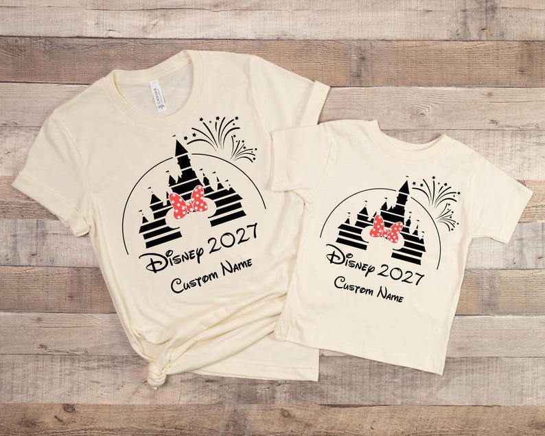 May include: Two cream-colored t-shirts with a black graphic of a castle, fireworks, and a red bow. The text "Disney 2027" and "Custom Name" are printed below the graphic. One shirt is adult size, the other is child size.