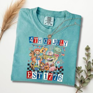 May include: A turquoise Comfort Colors t-shirt with a graphic of Toy Story characters celebrating the 4th of July. The text "4TH OF JULY" and "EST 1776" are in red, white, and blue. A silver necklace is draped over the shirt.