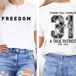 K&ouml;nnte beinhalten: Wei&szlig;es T-Shirt mit dem Wort "FREEDOM" in fetten schwarzen Buchstaben auf der Vorderseite. Auf der R&uuml;ckseite des Shirts steht der Text "THANK YOU, CHARLIE" &uuml;ber der Zahl "31" mit Bildern und dem Text "A TRUE PATRIOT 1993-2025".