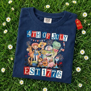 May include: A navy blue t-shirt with a graphic of cartoon characters and the text "4th of July EST 1776". The shirt has a crew neck and a "4XL" tag. The t-shirt is displayed on a bed of green grass.