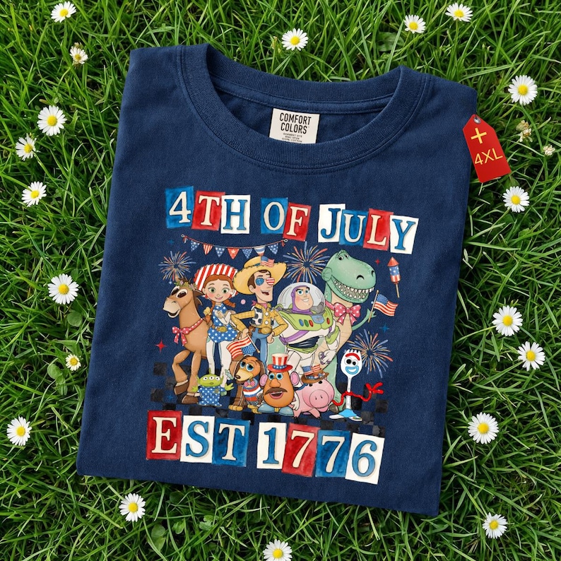 May include: A navy blue t-shirt with a graphic of cartoon characters and the text "4th of July EST 1776". The shirt has a crew neck and a "4XL" tag. The t-shirt is displayed on a bed of green grass.