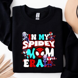 Puede incluir: Sudadera negra con un estampado gr&aacute;fico de personajes de Spider-Man y el texto "In My Spidey Mom Era".