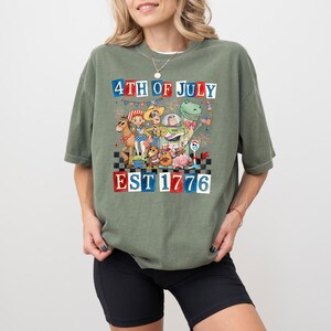May include: An olive green t-shirt with a vibrant graphic. The design features cartoon characters and the text "4TH OF JULY EST 1776". The graphic includes fireworks and a checkered pattern, ideal for celebrating Independence Day.