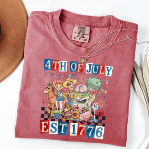 May include: A red t-shirt with a graphic featuring characters and the text "4th of July EST 1776". The shirt has a crew neck and short sleeves. A silver necklace with a pendant is draped across the shirt.