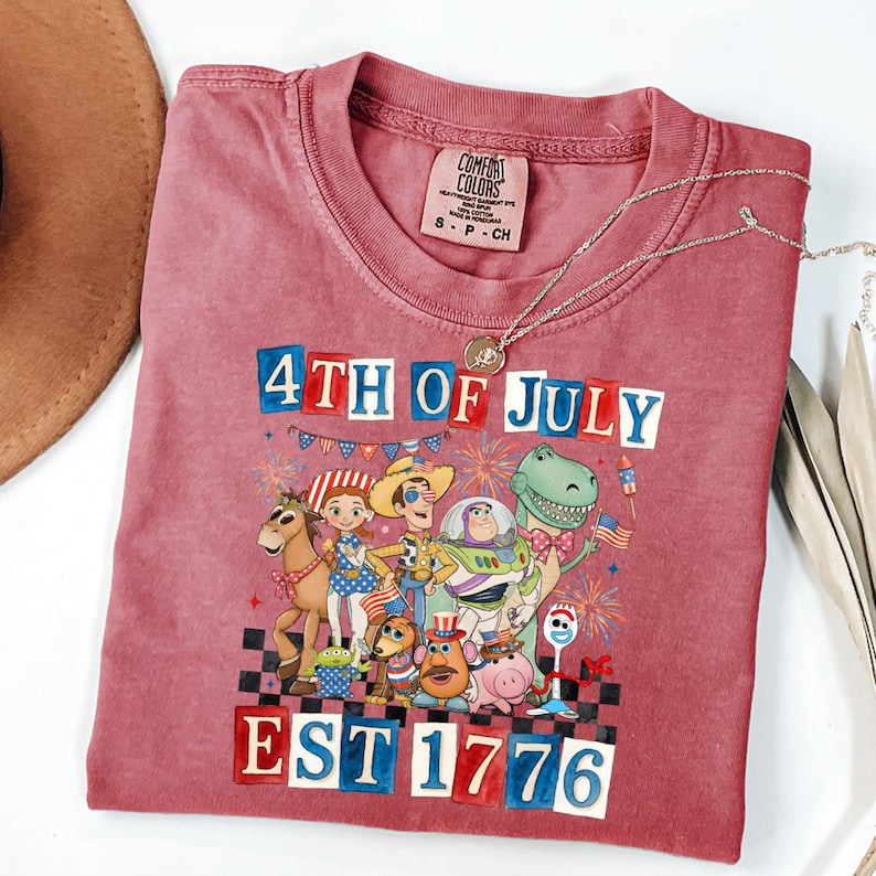 May include: A red t-shirt with a graphic featuring characters and the text "4th of July EST 1776". The shirt has a crew neck and short sleeves. A silver necklace with a pendant is draped across the shirt.