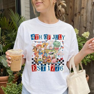 May include: White t-shirt with a colourful graphic that reads "4th of July EST 1776". The design includes cartoon characters and fireworks, with red, white, and blue details. A casual, festive design.