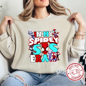 Puede incluir: Una sudadera beige claro con un estampado gr&aacute;fico de personajes de Spider-Man y el texto "In My Spidey Sis Era".