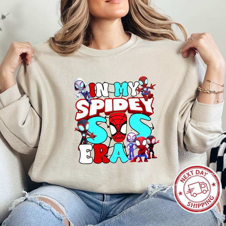 Puede incluir: Una sudadera beige claro con un estampado gr&aacute;fico de personajes de Spider-Man y el texto "In My Spidey Sis Era".