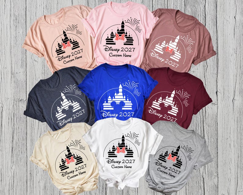 May include: A collection of various colored t-shirts featuring a castle design with fireworks, a bow, and the text "Disney 2027 Custom Name". The shirts are in shades of pink, blue, gray, and beige.