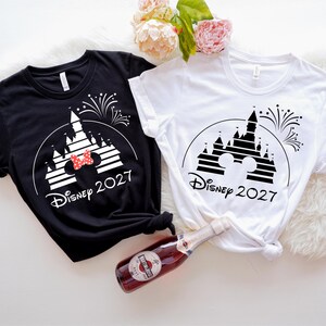 May include: Black and white t-shirts with a Disney castle graphic, fireworks, and the text "Disney 2027". One shirt has a red bow. A bottle of sparkling wine is in the image.