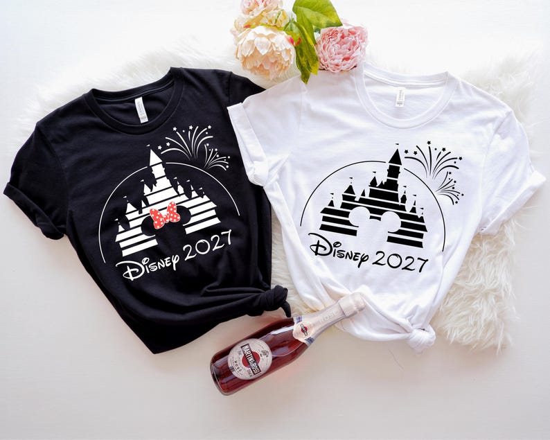 May include: Black and white t-shirts with a Disney castle graphic, fireworks, and the text "Disney 2027". One shirt has a red bow. A bottle of sparkling wine is in the image.