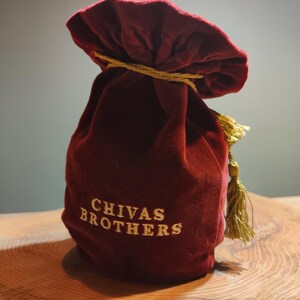 May include: A red velvet bag with a gold drawstring and tassel. The bag has the words "CHIVAS BROTHERS" embroidered on the front in gold thread.