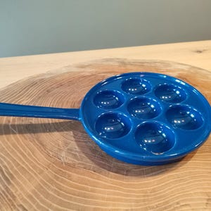 May include: A blue ceramic pan with a handle, designed for making individual portions of food. The pan has eight round indentations.