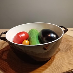 Könnte beinhalten: Eine weiße Keramikschüssel mit schwarzem Rand und Griffen, gefüllt mit verschiedenen Gemüsesorten, darunter eine rote Tomate, eine kleine orangefarbene Tomate, eine grüne Gurke und eine dunkelviolette Aubergine.