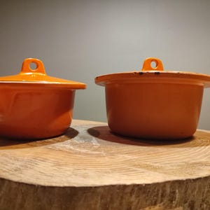 以下が含まれることがあります： ふた付きのオレンジ色のセラミック製のキャセロールが2つ。小さい方は手前に、大きい方は奥に置かれています。どちらも木製の台の上に置かれています。