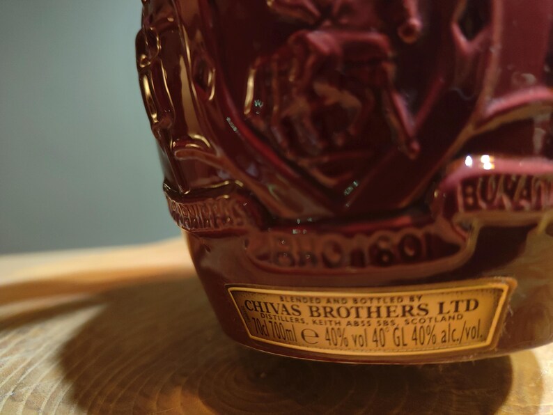 May include: A dark red bottle with a gold label that reads "BLENDED AND BOTTLED BY CHIVAS BROTHERS LTD DISTILLERS, KEITH ABSS SNS. SCOTLAND d 700ml e 40% vol 40&deg;GL 40% alc./vol."