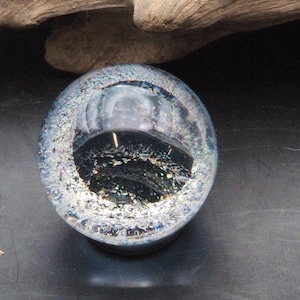 May include: A clear glass sphere with a dark interior and a textured, iridescent exterior. The sphere sits on a dark surface, reflecting light and creating a shimmering effect. The sphere is a decorative object.