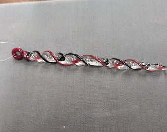 May include: A clear glass swirl with red and black detailing. The decorative item has a loop at one end and tapers to a point. A thin, clear string is attached to the loop. The glass piece is on a grey surface.