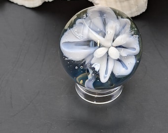 May include: A clear glass paperweight with a white and light blue floral design. The paperweight is round and sits on a clear, circular base. Air bubbles are visible within the glass, and a large seashell is in the background.