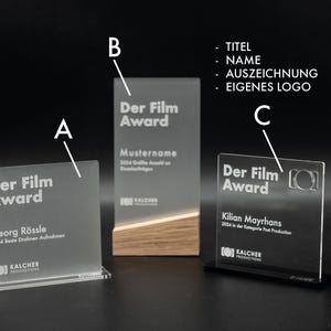 Può includere: Tre targhe premio in acrilico trasparente con un designo inciso in bianco. Le targhe presentano il testo "Der Film Award" e i nomi dei premiati. Le targhe sono su basi nere e in legno.