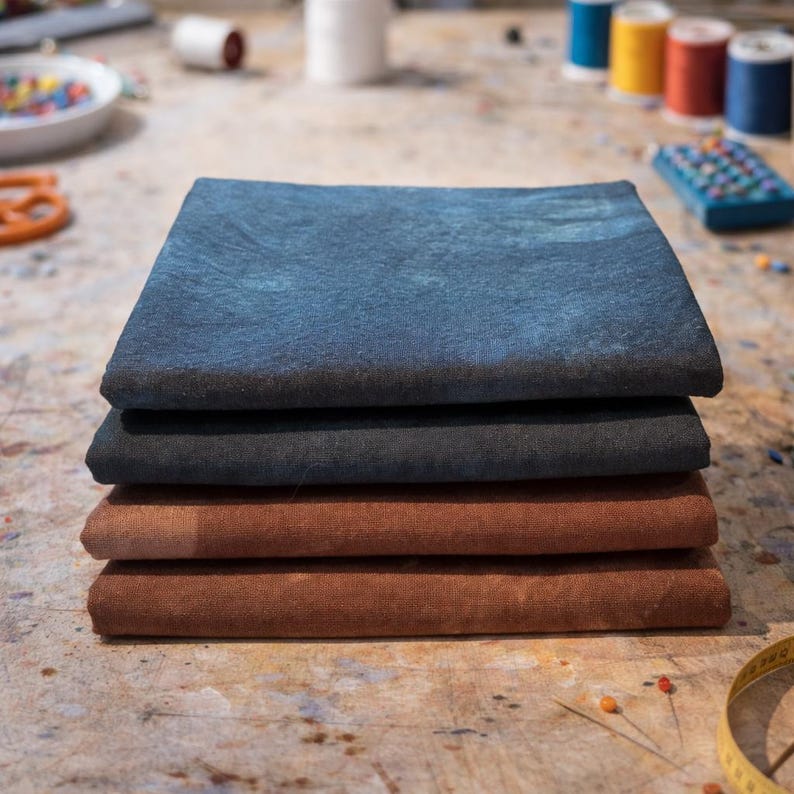 May include: A stack of four folded fabric squares in shades of blue and brown. The top square is a mottled blue, followed by a dark gray, then two shades of brown. The fabrics are stacked on a textured surface, suggesting a craft or sewing project.