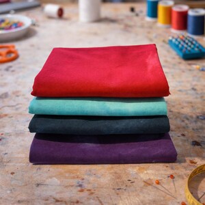 Può includere: Una pila di quattro quadrati di tessuto piegati in colori vivaci: rosso, turchese, verde scuro e viola. I tessuti sono impilati su una superficie di legno, con forniture di cucito sullo sfondo, suggerendo un tema di cucito.