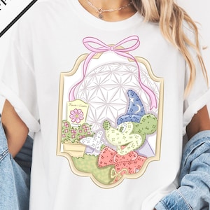 A woman wearing a white t-shirt with a colorful graphic design of a space station and various objects, including a flower and a cactus, is standing against a white background.