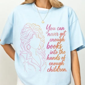 May include: A light blue t-shirt featuring a line art illustration of a person with voluminous hair and the text, "You can never get enough books into the hands of enough children," printed in a gradient of pink, orange, and yellow hues.