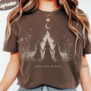 A woman wearing a brown t-shirt with a graphic design of three silhouetted figures and the text "girls will be girls" printed on it.