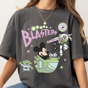 Pu&ograve; includere: T-shirt grigio scuro con una grafica a cartoni animati con Topolino, Buzz Lightyear e un alieno, con la scritta "Astro Blasters". Il design utilizza colori pastello e accenti di stelle.