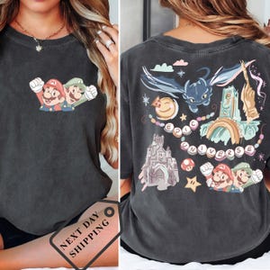 May include: Dark gray t-shirt featuring a colorful graphic of Mario and Luigi on the front. The back of the shirt displays a whimsical design with a castle, a dragon, and the words "EPIC UNIVERSE".