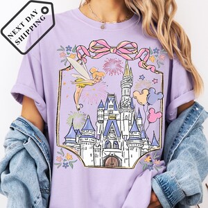 A person wearing a purple t-shirt with a castle and fairy graphic printed on it, along with a denim jacket.