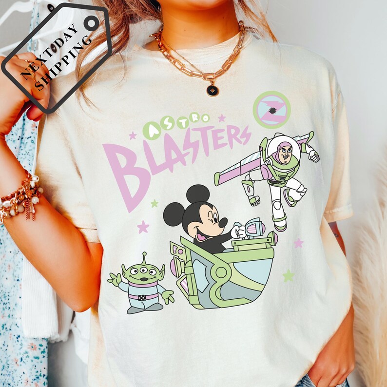 Pu&ograve; includere: T-shirt color crema con una grafica colorata di Topolino, Buzz Lightyear e un alieno di Toy Story. Il testo "Astro Blasters" &egrave; in rosa e verde. La maglietta ha una vestibilit&agrave; ampia.