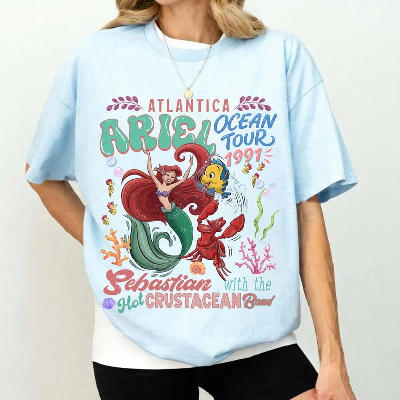 A person wearing a light blue t-shirt with a colorful graphic design featuring a mermaid character and text related to the Ariel Ocean Tour.