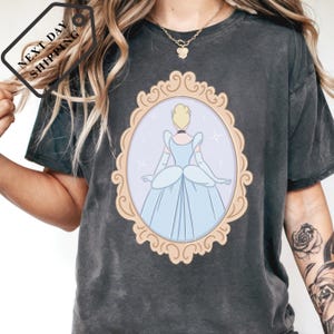 A person wearing a black t-shirt with a graphic of a woman in a blue dress, framed by an ornate golden border.