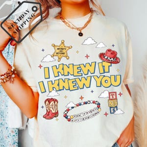 A person wearing a white t-shirt with a graphic design featuring a cowboy hat, clouds, and stars, along with the text "I KNEW IT I KNEW YOU" and other cartoon-style elements.