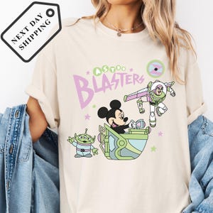 Pu&ograve; includere: T-shirt color crema con un design a cartoni animati con la scritta "Astro Blasters". Il design include Topolino, Buzz Lightyear e un alieno verde. La maglietta ha un girocollo e maniche corte.