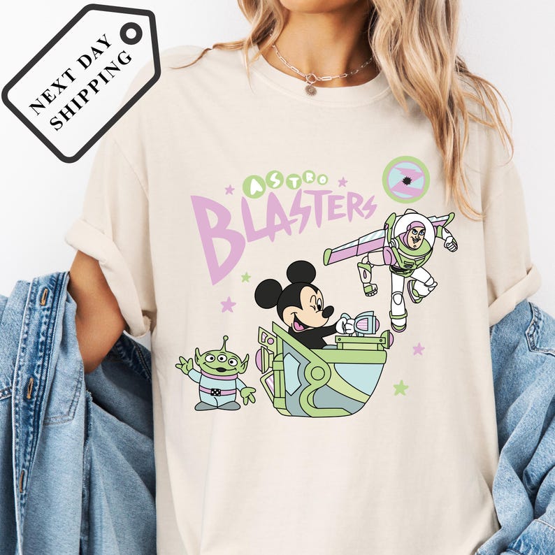 Pu&ograve; includere: T-shirt color crema con un design a cartoni animati con la scritta "Astro Blasters". Il design include Topolino, Buzz Lightyear e un alieno verde. La maglietta ha un girocollo e maniche corte.