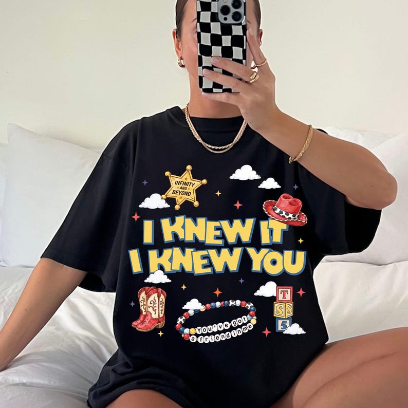 A person is sitting on a bed and taking a selfie with a black t-shirt featuring a graphic design.