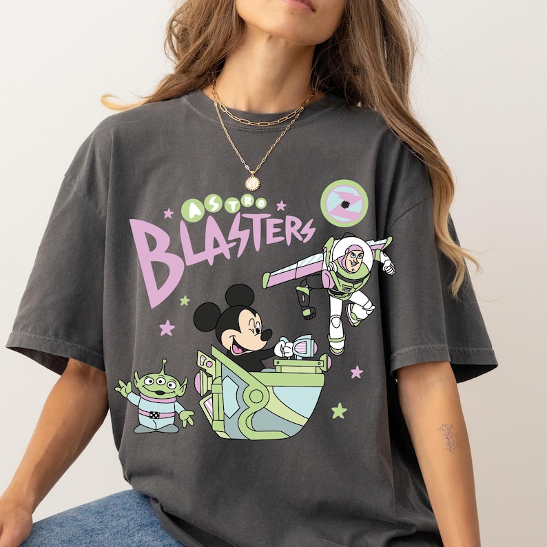 Pu&ograve; includere: T-shirt grigio scuro con una grafica a cartoni animati con Topolino, Buzz Lightyear e un alieno, con la scritta "Astro Blasters". Il design utilizza colori pastello e accenti di stelle.