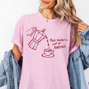 A woman wearing a pink t-shirt with a graphic design of a coffee pot pouring coffee into a cup, accompanied by the text "Pour yourself a cup of ambition".